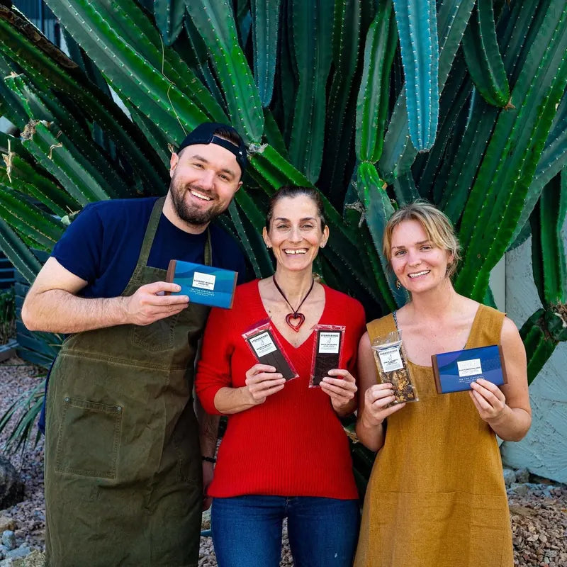Three of the Stone Grindz crew smiling in front of a tall cactus, each holding chocolate bars